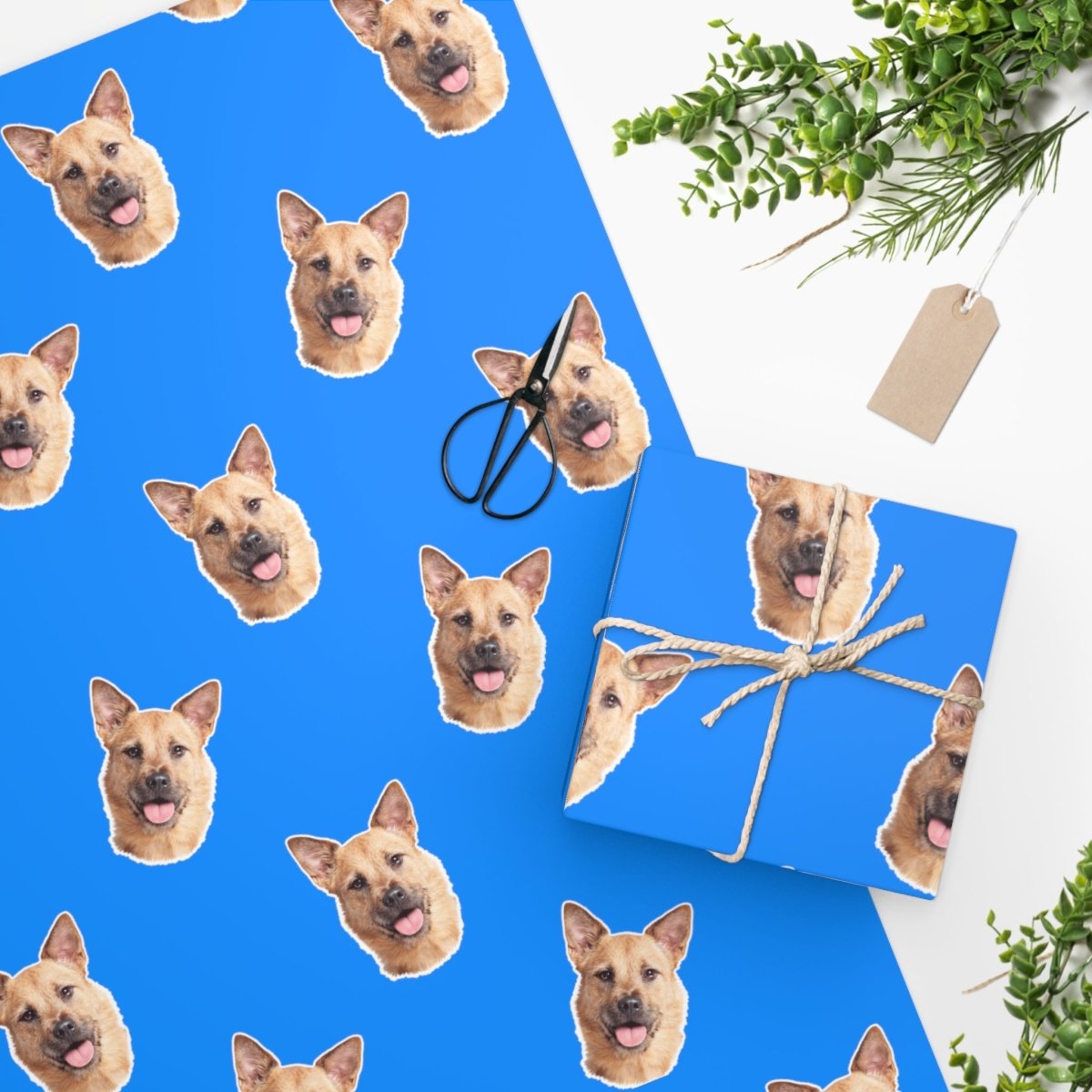 Wrapping paper with a repeated pattern of a dog's face on a blue background, placed next to a blue gift box with a rope handle and a tag.